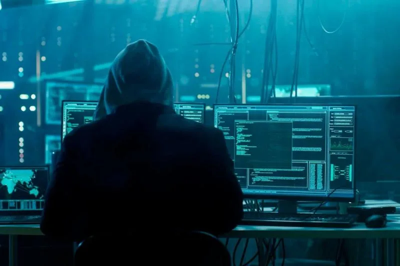 Hooded figure seated at a multi-monitor workstation displaying code in a dim, blue-toned room