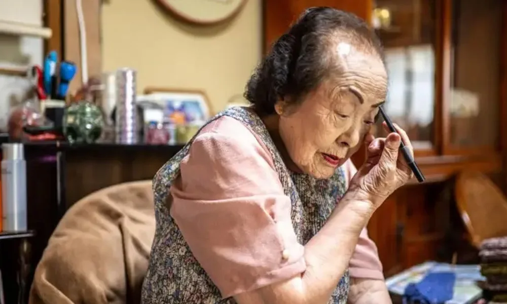 Elderly woman sitting at a cluttered table in a warm home, applying eye makeup with a brush to her eye.