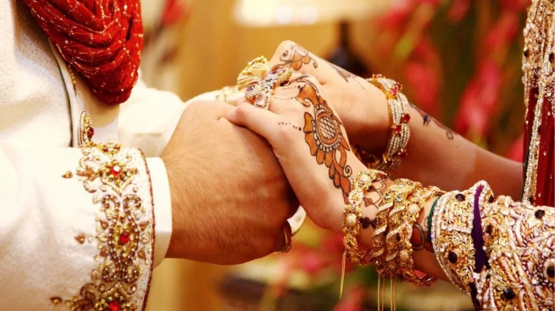 Bride and groom join hands in a ceremonial pose, adorned with henna and gold jewelry in ornate wedding attire