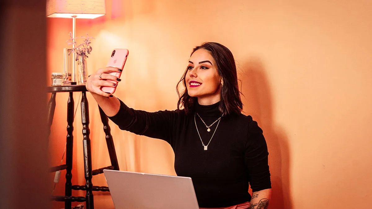 Woman in a black turtleneck takes a selfie with a pink smartphone, laptop open in front in a warm, orange-lit room.