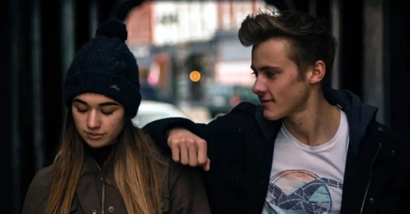 Two young people walk side by side on a city street; the man has his arm around the woman's shoulder as they look down and smile slightly.