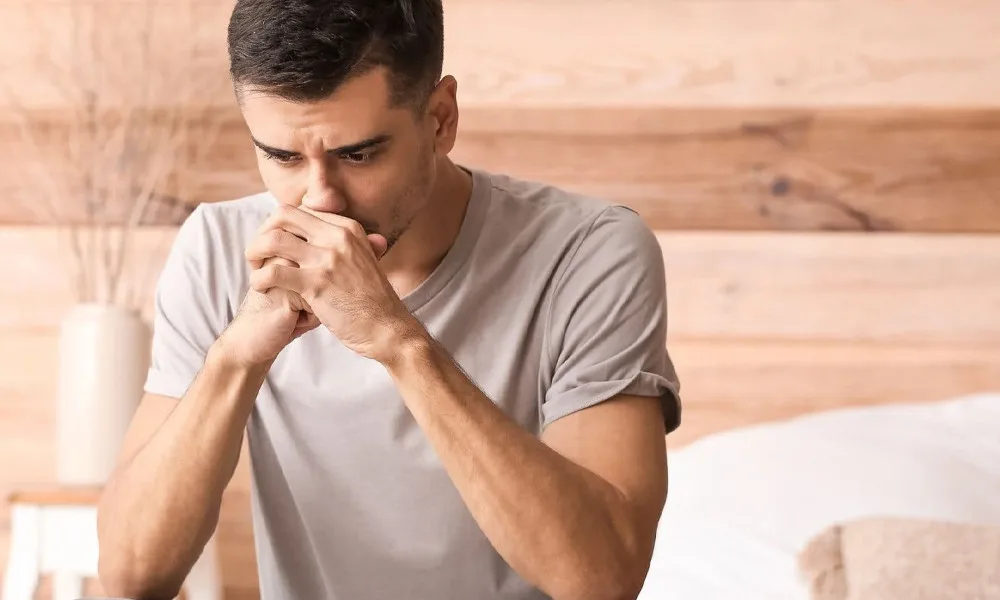 Man in a gray t-shirt sits indoors, hands clasped under his chin, looking down in thought/concerned.
