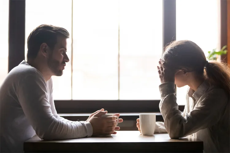 Man and woman sit across from each other at a table by a large window, having a serious conversation; a white mug sits between them.