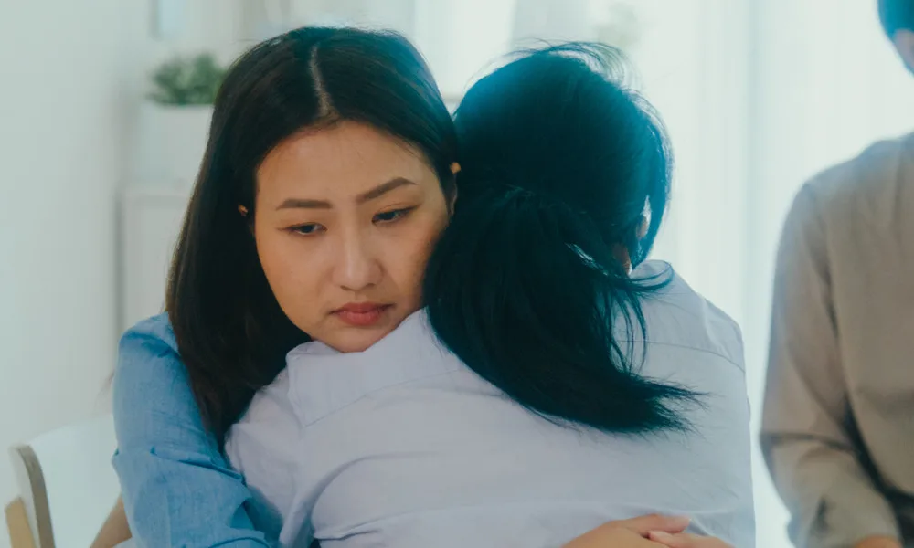 Two women hug closely in a bright room, one resting her head on the other's shoulder with a calm, somber expression on her face.