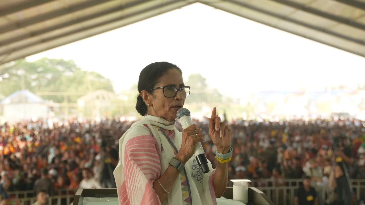 Woman wearing glasses on stage speaking into a microphone to a large outdoor crowd.