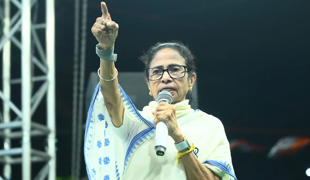 Senior woman with glasses speaks into a microphone on stage, raising her right hand with index finger pointing upward.