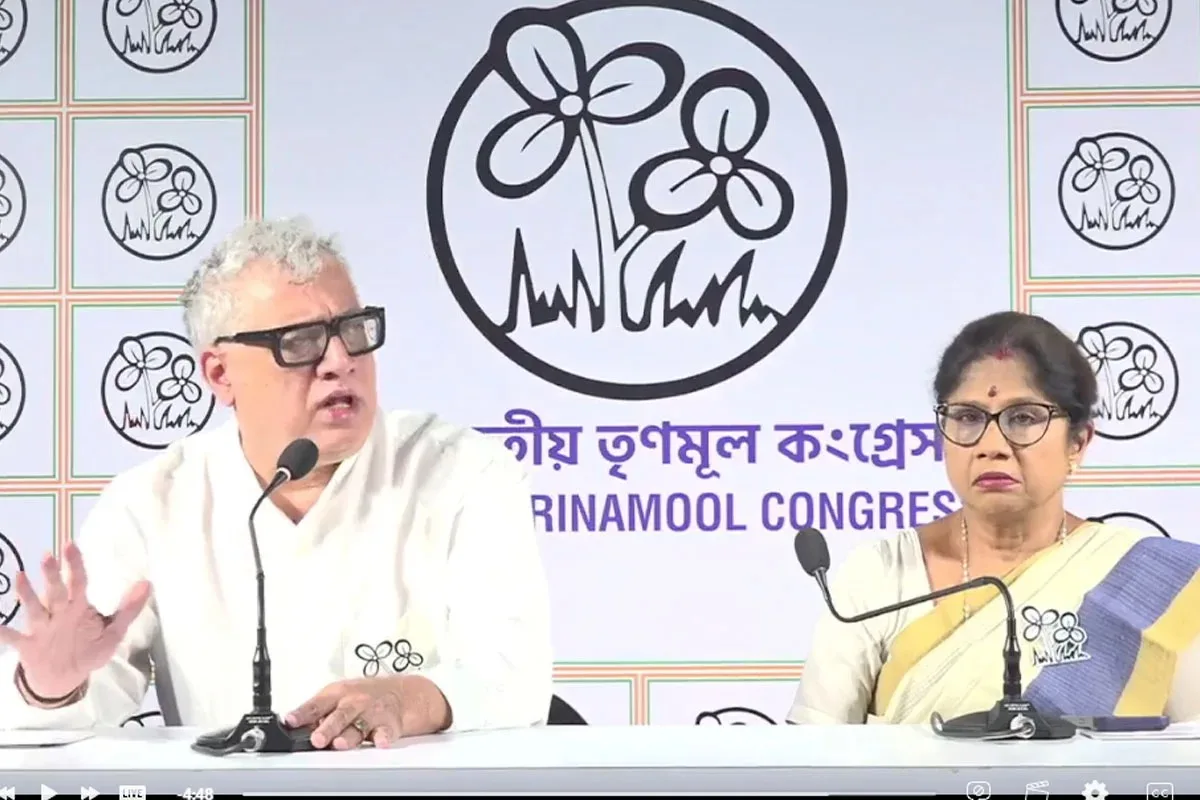 Two Indian politicians sit at a press conference desk with microphones, backdrop displaying a party logo and 'Congress' text in the background.