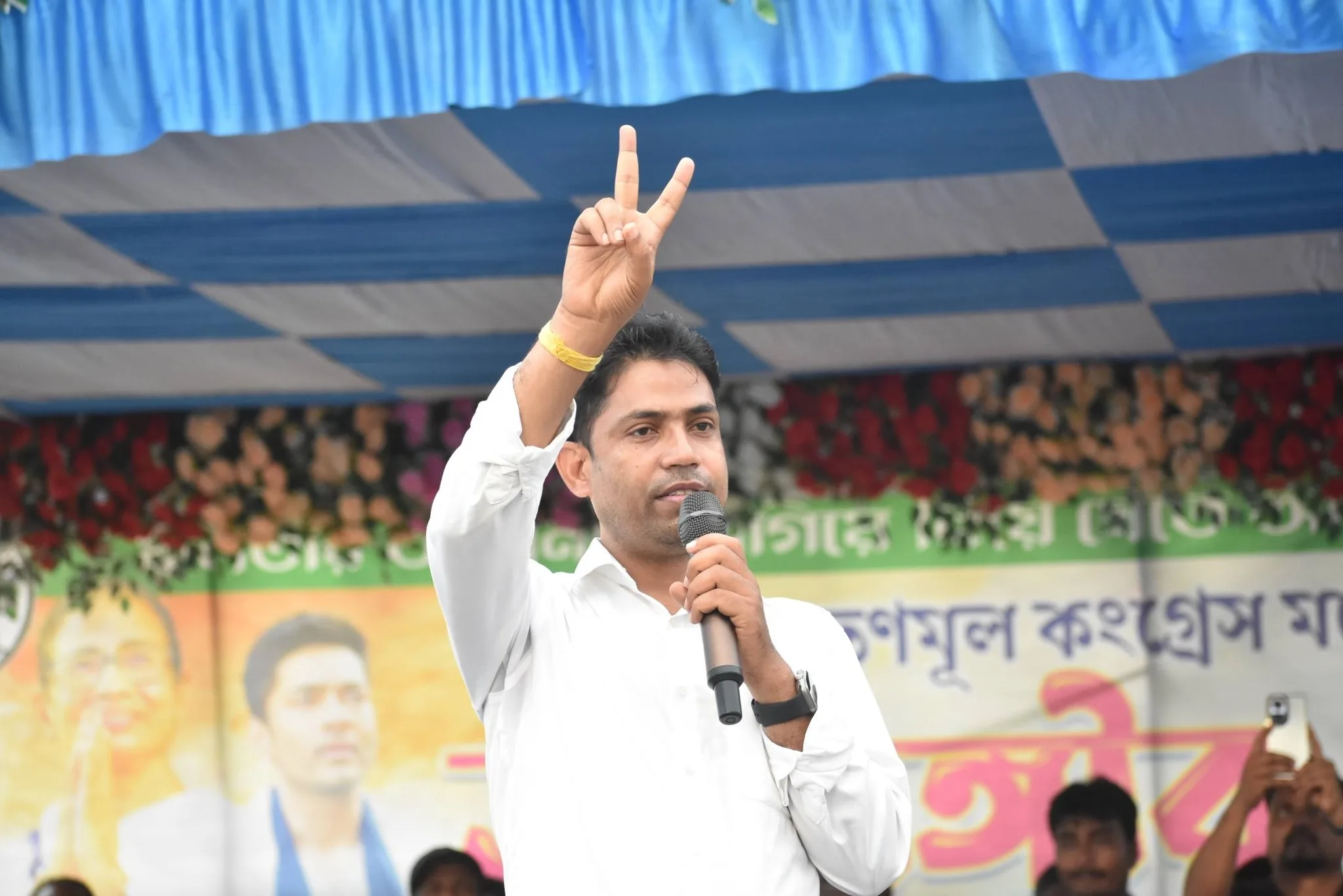 Man in white shirt on stage raises two fingers in a peace sign while speaking into a microphone at a rally.