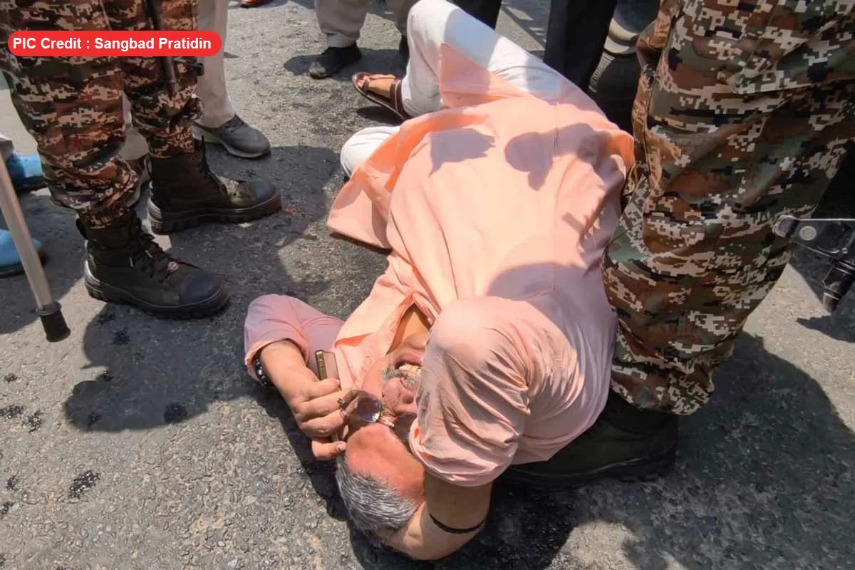 Man in a peach shirt lying on the road while surrounded by soldiers, one nurse or bystander crouching nearby, suggesting an injured person being attended to