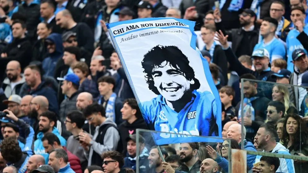 Fans at a soccer match hold a blue banner featuring a smiling portrait of a man amid a crowded stadium.