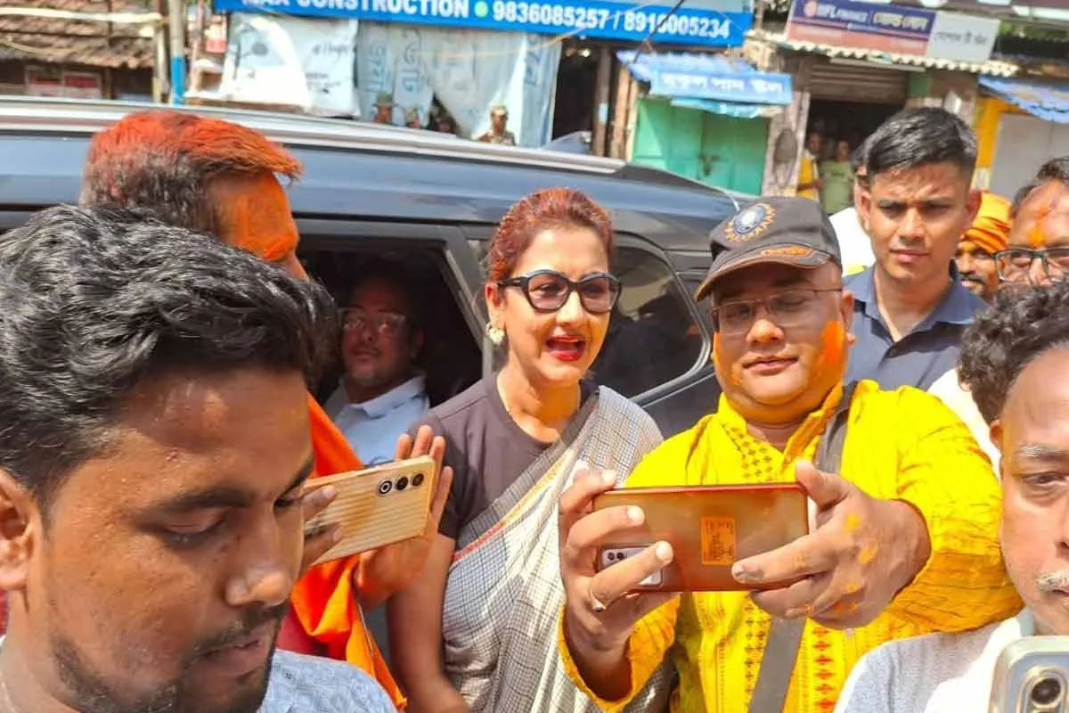 Group of people taking a selfie in a busy street; a woman wearing sunglasses and a saree smiles beside a man in a bright yellow scarf, all posing with smartphones outstretched.