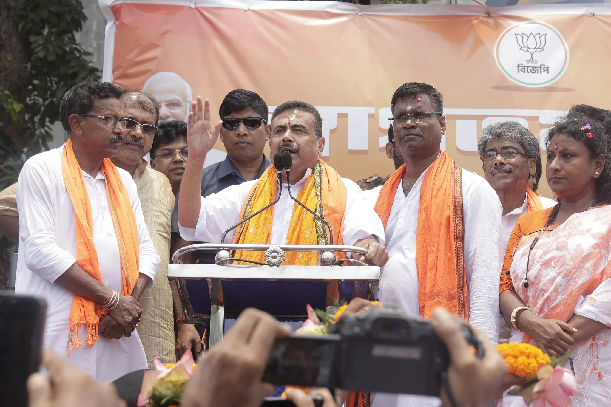 Man at a podium delivering a speech, surrounded by supporters in white clothing and orange scarves at a rally with a banner behind him.
