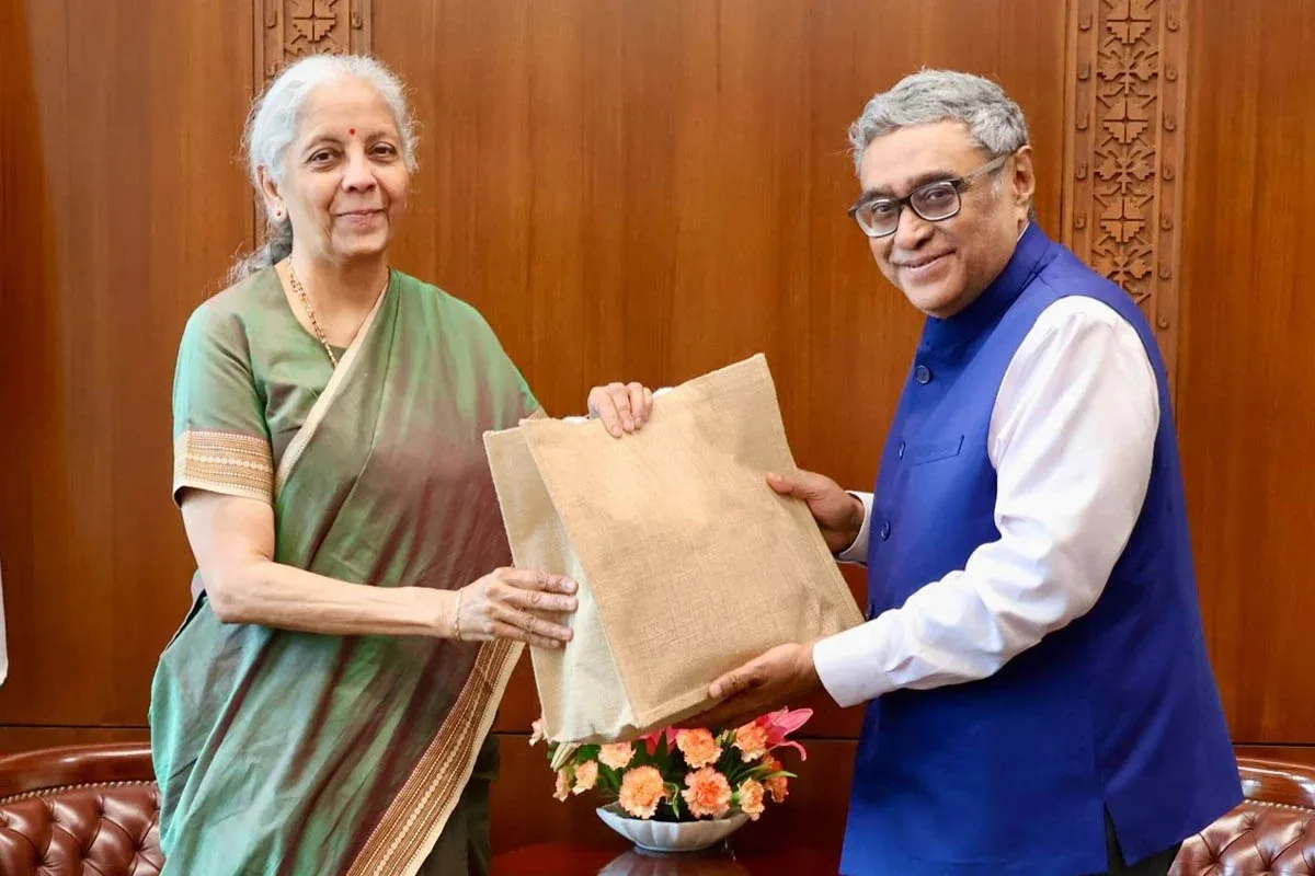 Two Indian officials exchange a burlap gift bag in a formal room with wood-paneled walls and a flower arrangement nearby.