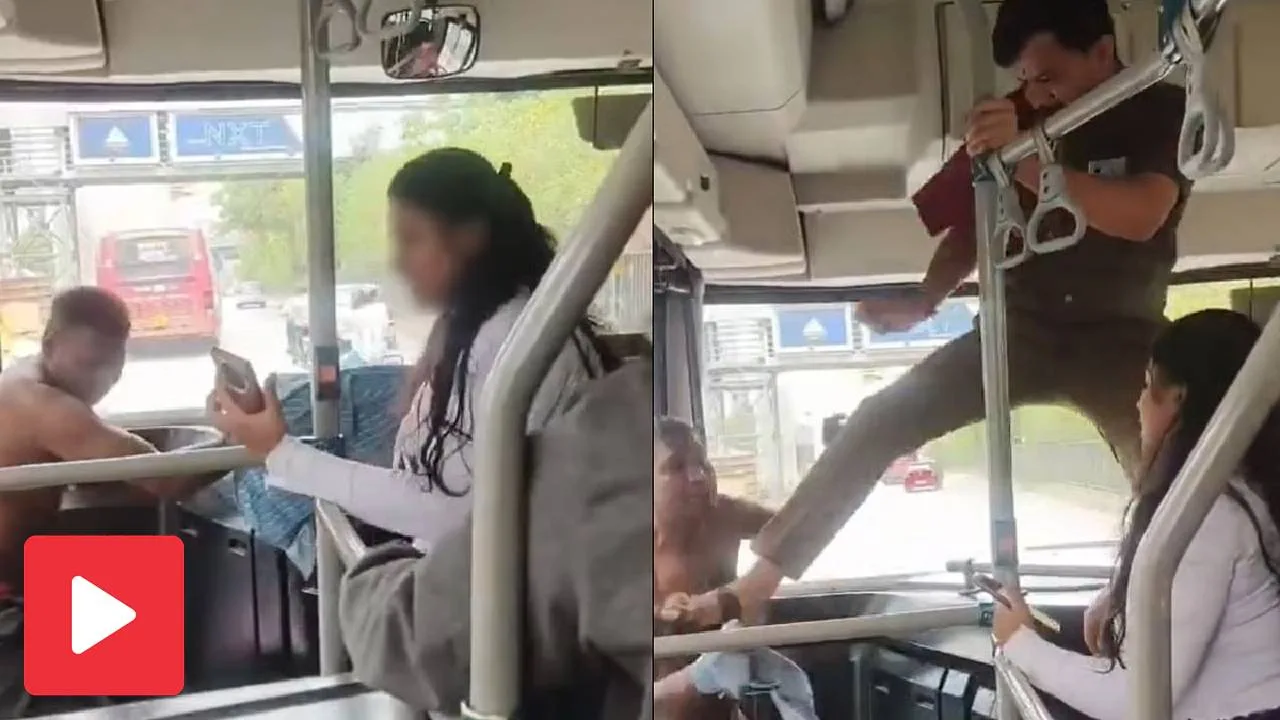 Inside a city bus, a shirtless man sits near the driver while a woman in a white coat stands holding a phone and looking at him; bus interior with poles and straps visible.