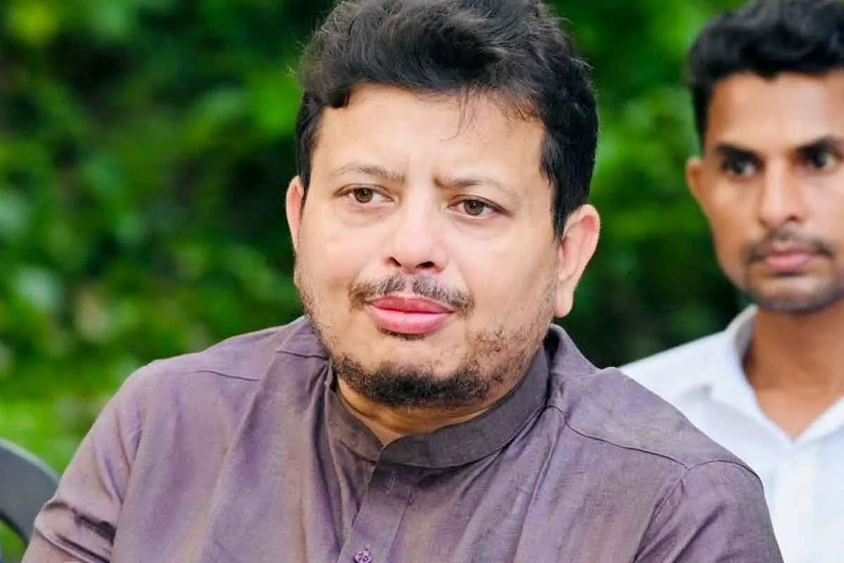 Close-up of a man with short dark hair and a beard wearing a purple kurta, outdoors with greenery behind him.