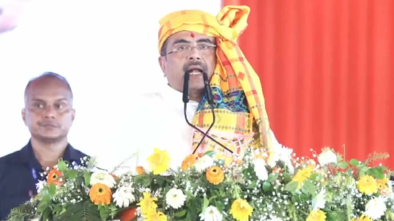 Man in a yellow patterned turban speaks at a flower-decked podium, with a man in a dark shirt behind him.